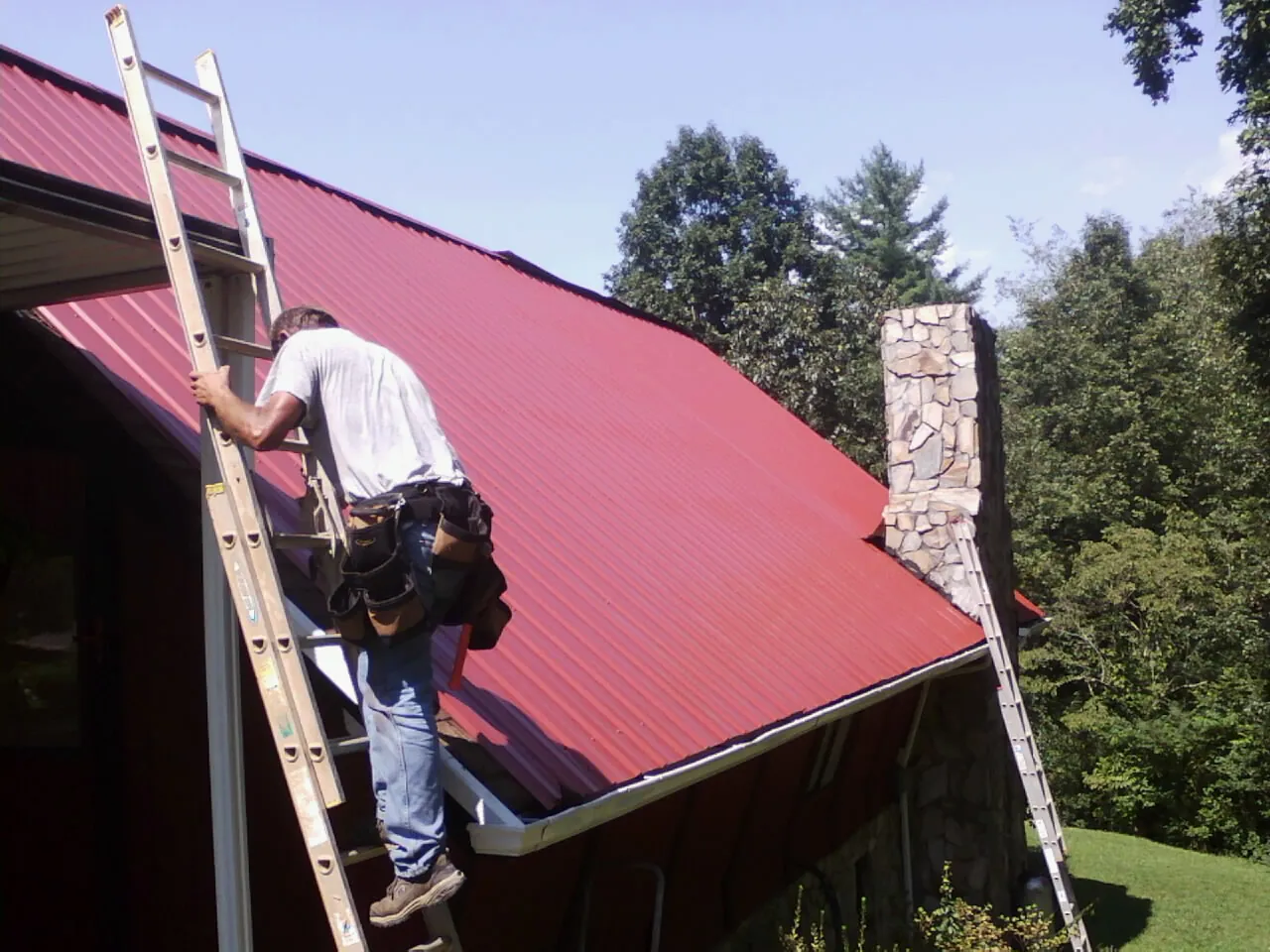 Roofer on a ladder beside a red metal roof and stone chimney — Soffit & Fascia Repair by Roofing Brothers in Anatone